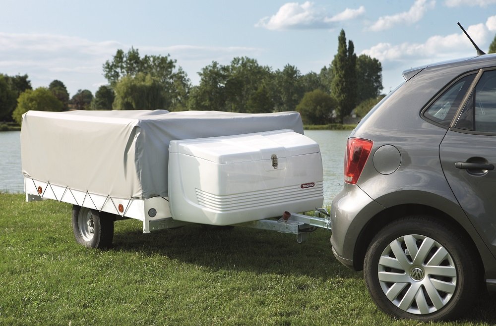 A gray car towing a Camplair trailer by a lake with trees and blue sky in the background.