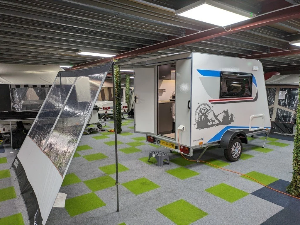 A compact white camper trailer with a side door open, parked indoors on a green and gray checkered carpet, with a black solar panel leaning against a pole and an awning extended, featuring mountain and forest graphics.