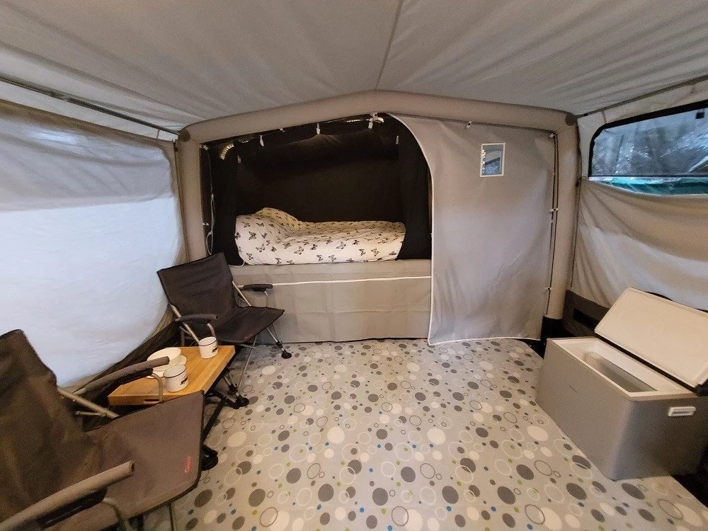 Interior of a Campmaster Air with a bed, two chairs, a small table with cups, and a cooler, with patterned flooring and windows.