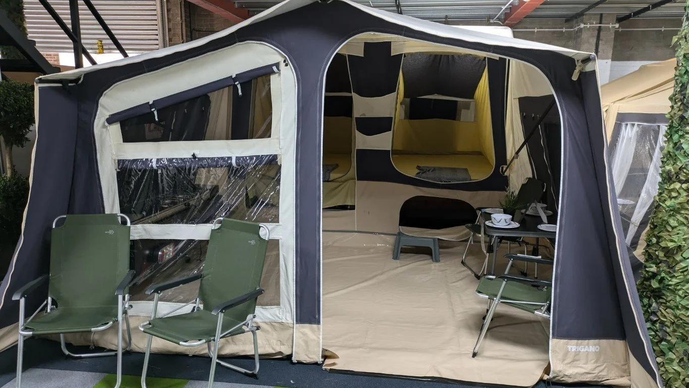 A large outdoor tent with a black and beige color scheme set up on a patio. The tent features clear plastic windows and an open front entrance. Inside, there are two green camping chairs, a black chair, a small table with cups, and some small potted plants. The background shows additional tents and metal stairs.