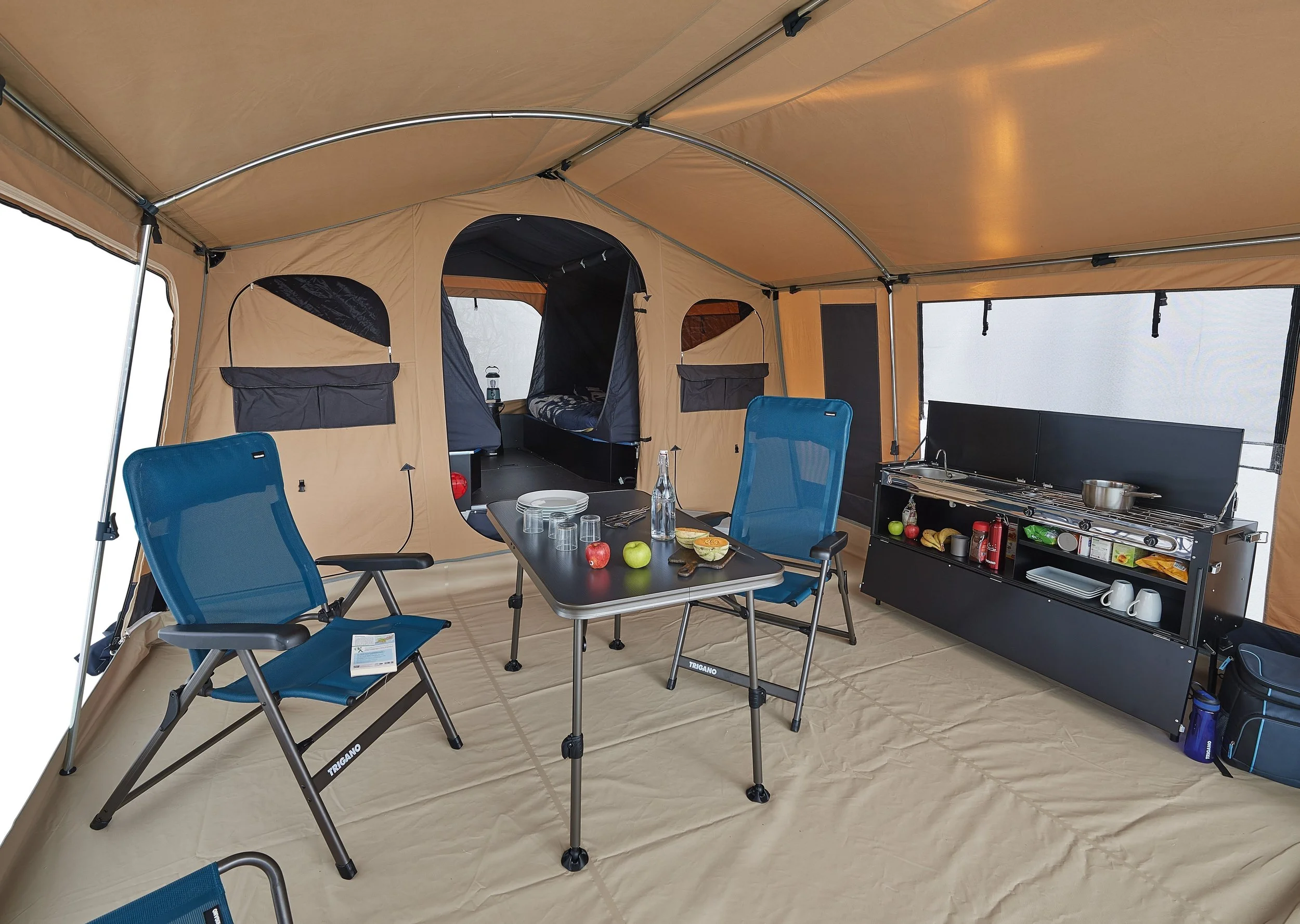 Inside a large camping tent with a kitchen area, table, and chairs. The table has apples, a plate of food, glasses, and a water bottle. Chairs are blue, and the kitchen includes a stove with utensils, cups, and food items.