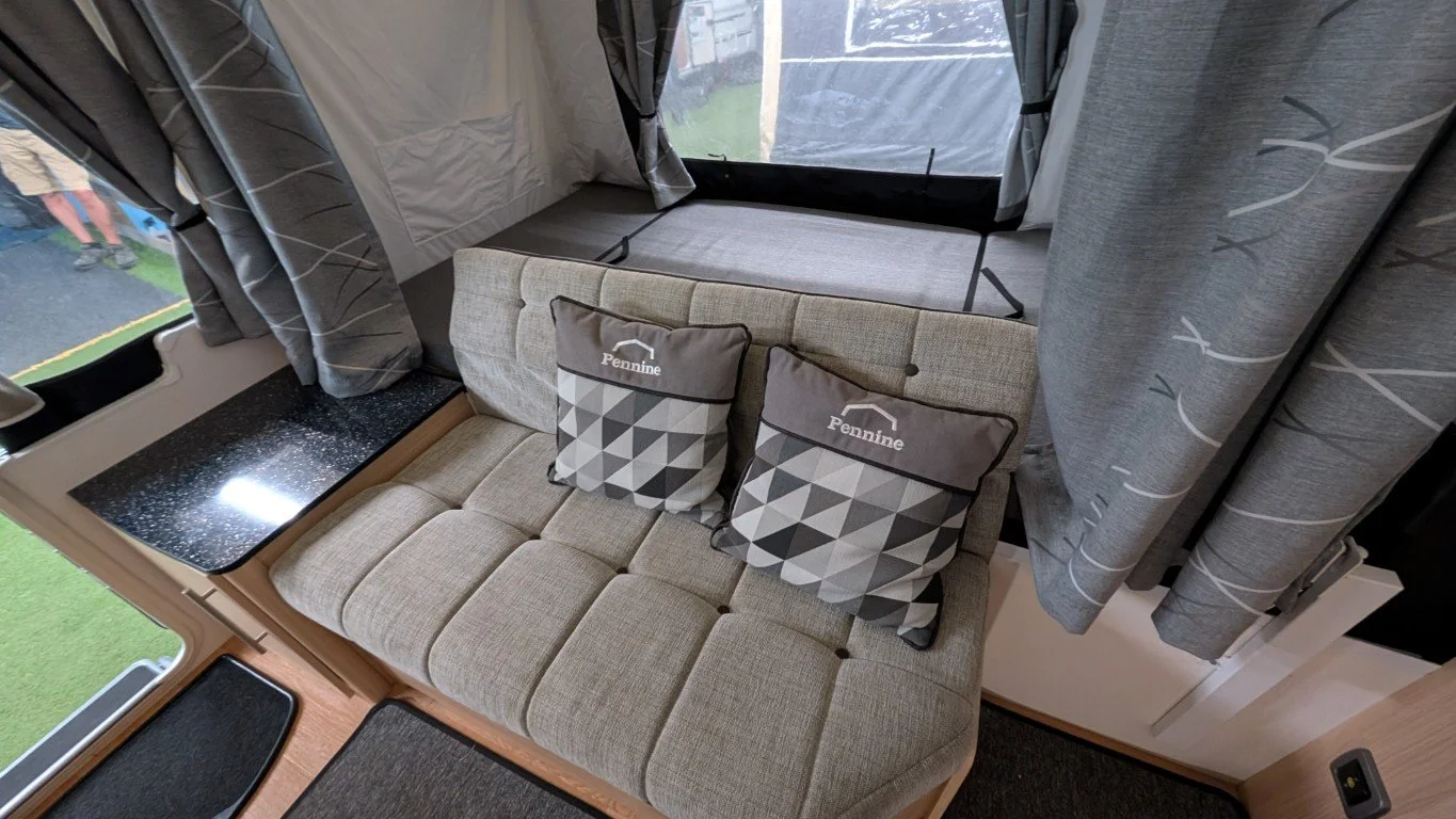 Interior of a camper or RV with a beige cushioned seating area, two decorative pillows with geometric patterns and the word 'Pennine', black window shades with a leaf pattern, and a window ledge with a black speckled surface.