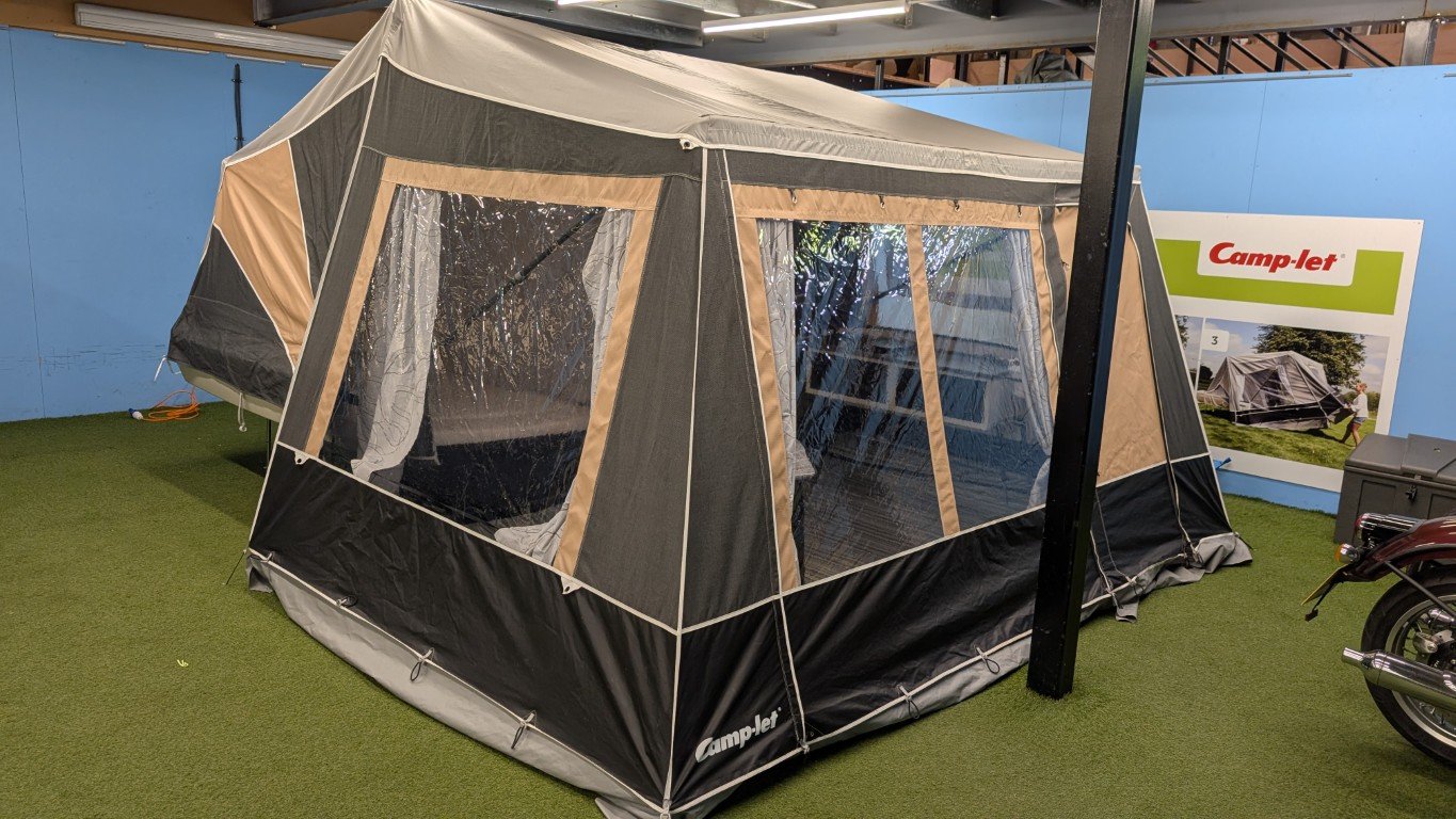 A large camping tent with beige and black fabric, set up indoors on artificial grass. The tent features multiple windows with clear plastic panels and beige curtains. A Camp-let promotional poster is visible in the background.