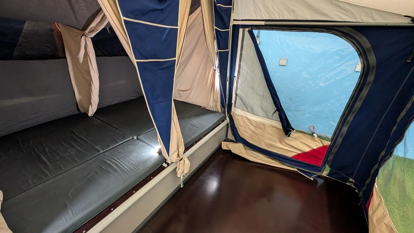 Interior of a camping tent with a bunk bed and a floor space, with a blue and beige color scheme and a window showing green scenery outside.