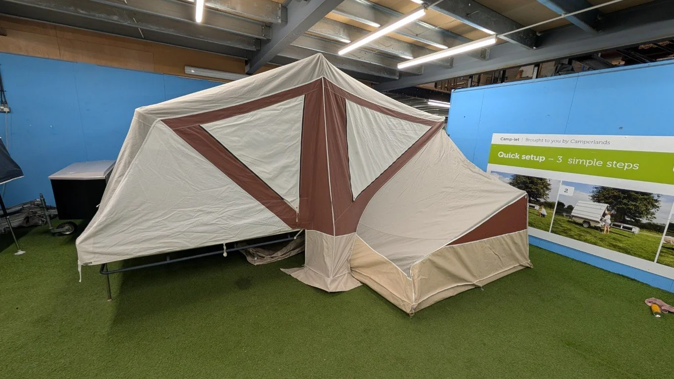 A beige and brown camping tent displayed indoors on artificial grass, with a blue wall and a sign describing quick setup steps in the background.