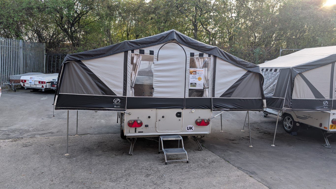 Camping trailer with extended awning setup, parked on paved surface, surrounded by other similar trailers, with trees in the background.