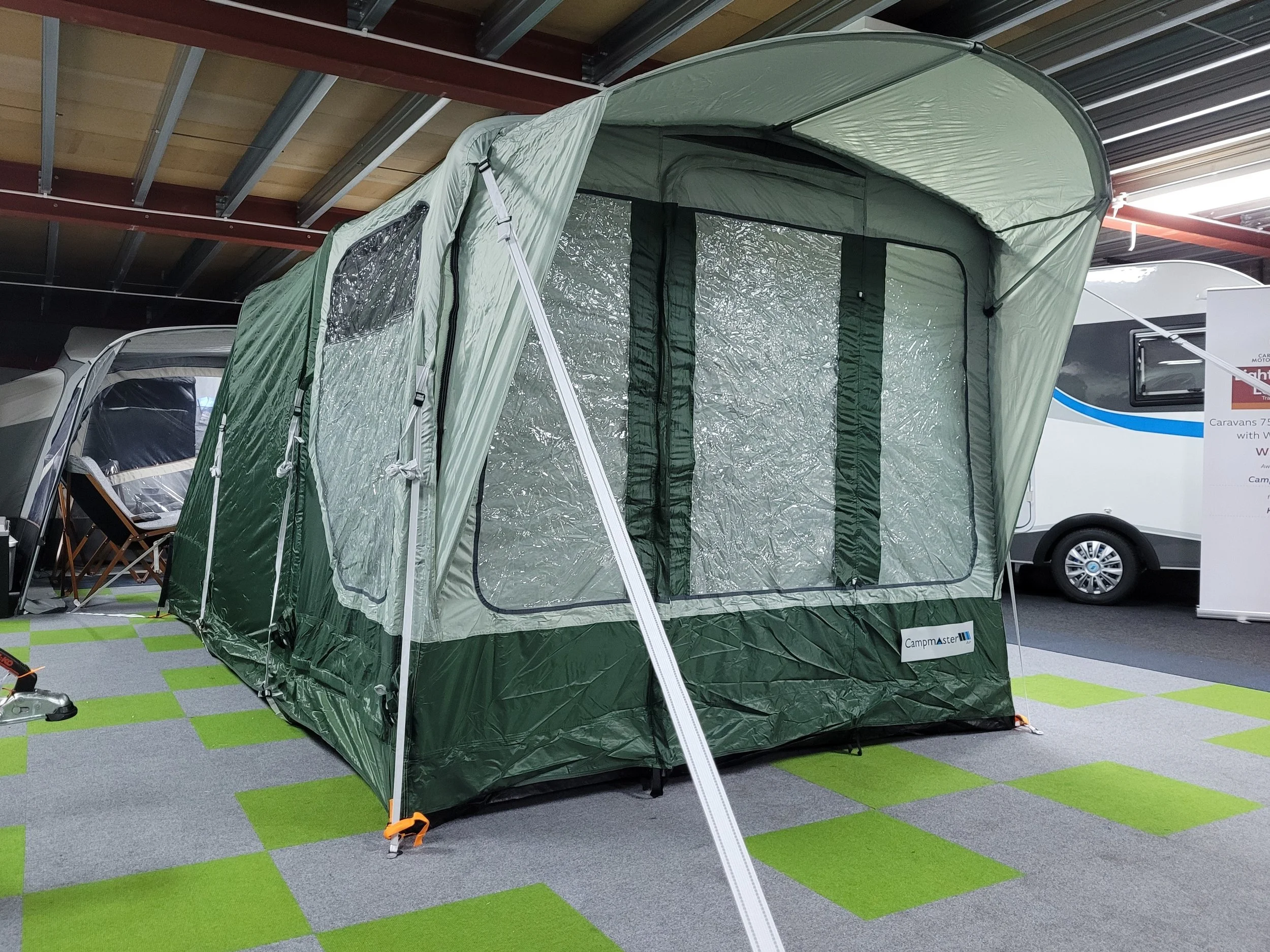 Campmaster Air A green and gray camping tent with a large zippered door, set up indoors on gray and green carpet, with other camping gear and caravans nearby.