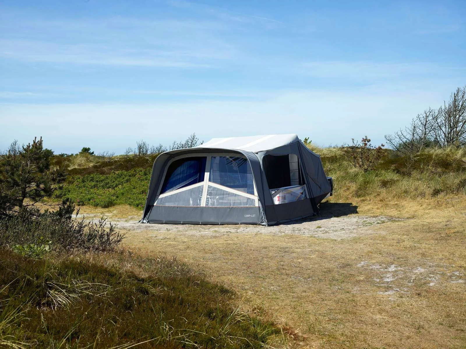 A black and white camping tent set up on a grassy field with a dirt patch, surrounded by sparse trees and bushes under a partly cloudy blue sky.