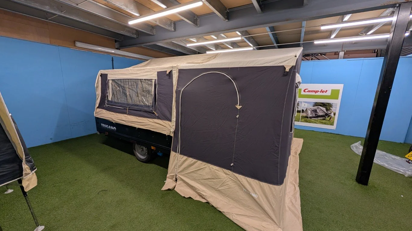 A compact tent with beige and dark gray fabric is mounted on a small trailer with a single wheel, displayed indoors on artificial grass. There is a sign in the background showing the tent on a grassy area.
