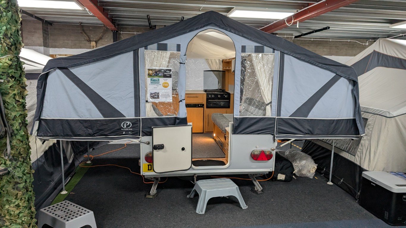 Small camping trailer with an extended canopy, inside is a kitchenette and seating area, set up indoors in a showroom or storage space.