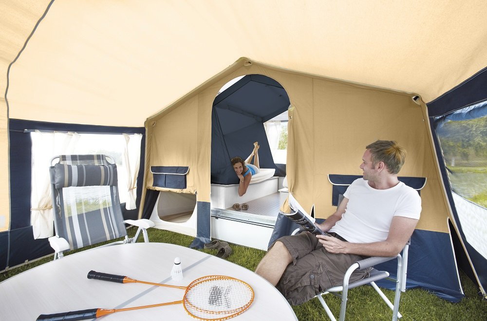 A family inside a camping tent, with a child lying on a bed in a smaller inner tent and a man sitting outside in a camping chair.