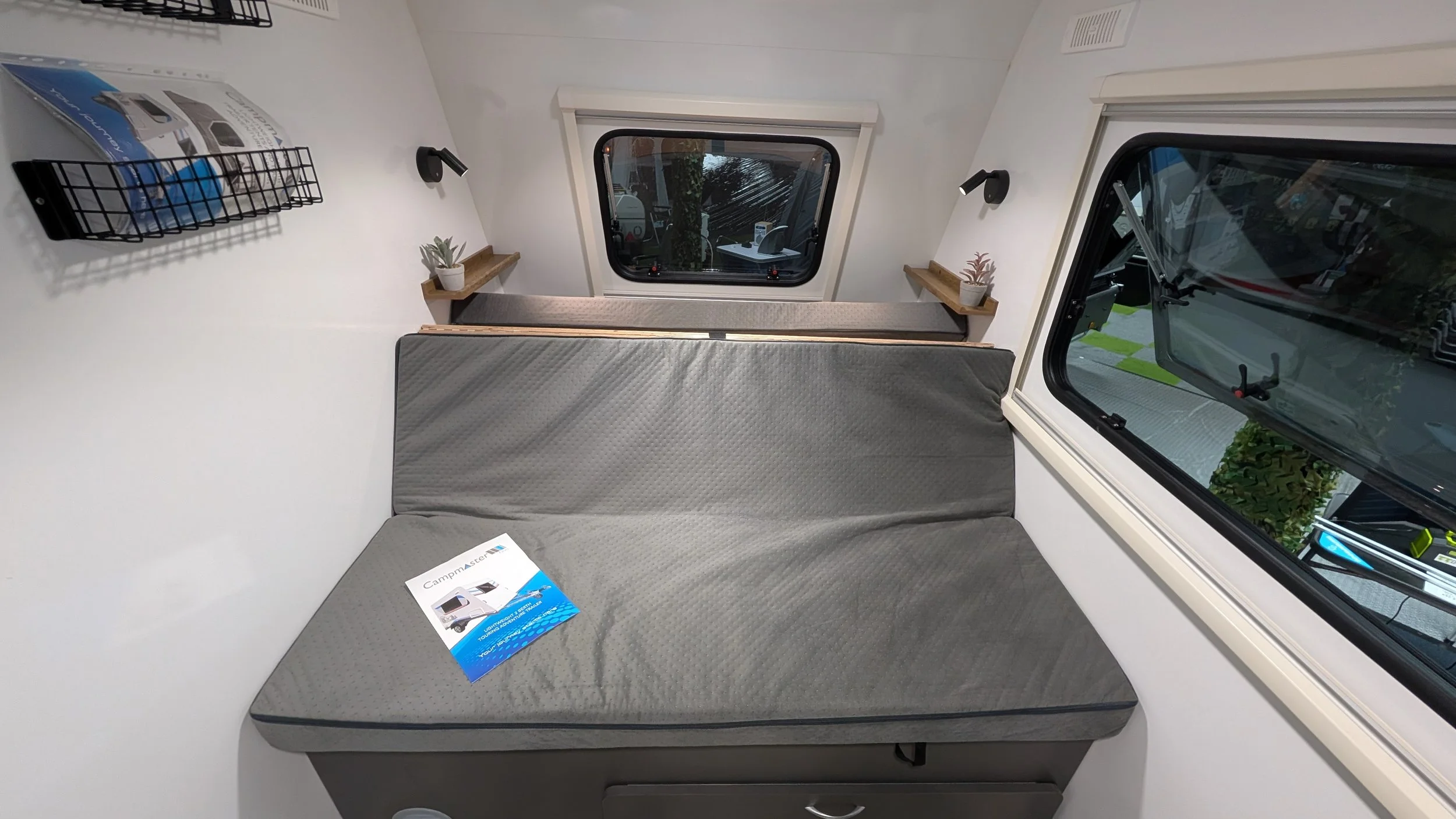 Interior of a small camper with a gray seating area, two small potted plants on wooden shelves, windows showing outside scenery, black wall-mounted lights, and a black wire basket on the wall, with a brochure on the seat.