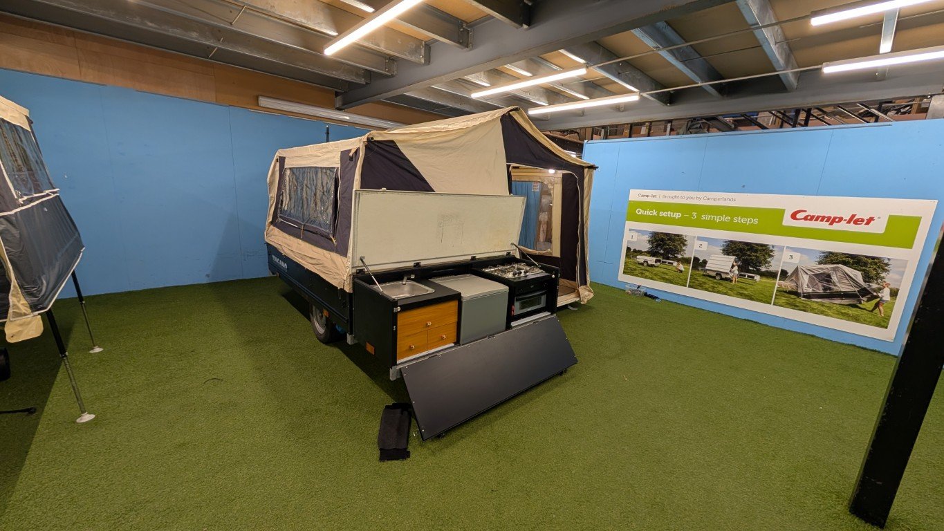 Display of a compact camping trailer with a built-in kitchen, set up indoors with artificial grass flooring and a blue wall, showing a step-by-step guide for quick setup.