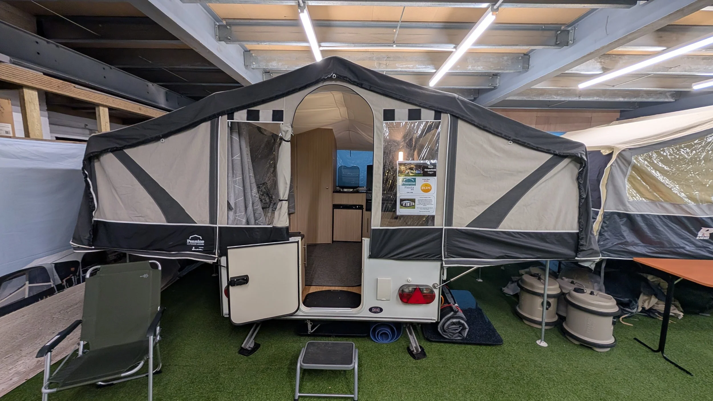 A camper trailer with a pop-up tent set up inside a store. The interior shows a kitchenette with a wooden cabinet and a small TV. Surrounding the trailer are camping chairs, a small table, and various camping gear, all on a grassy floor display.