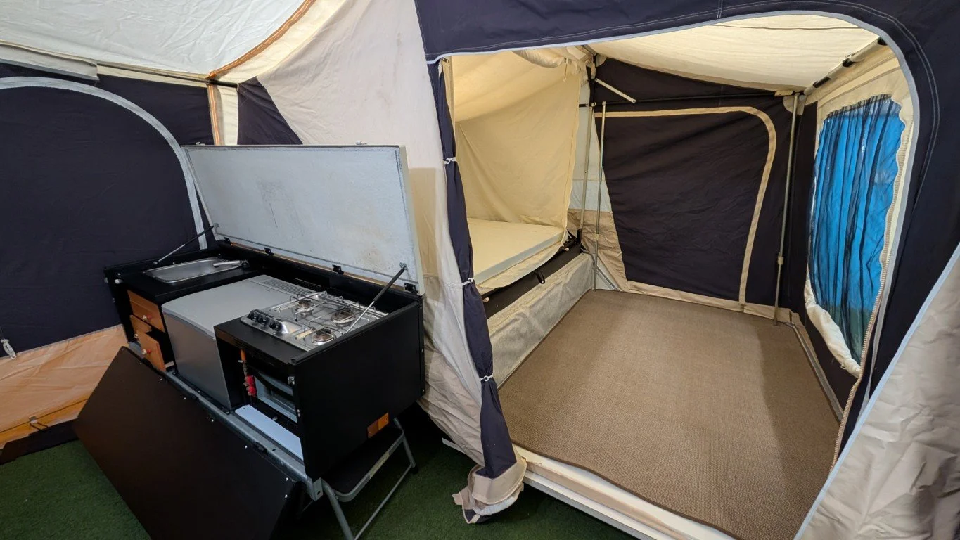 Inside a campsite tent with a sleeping cot, a small kitchenette with a stove and sink, and a beige carpeted floor.