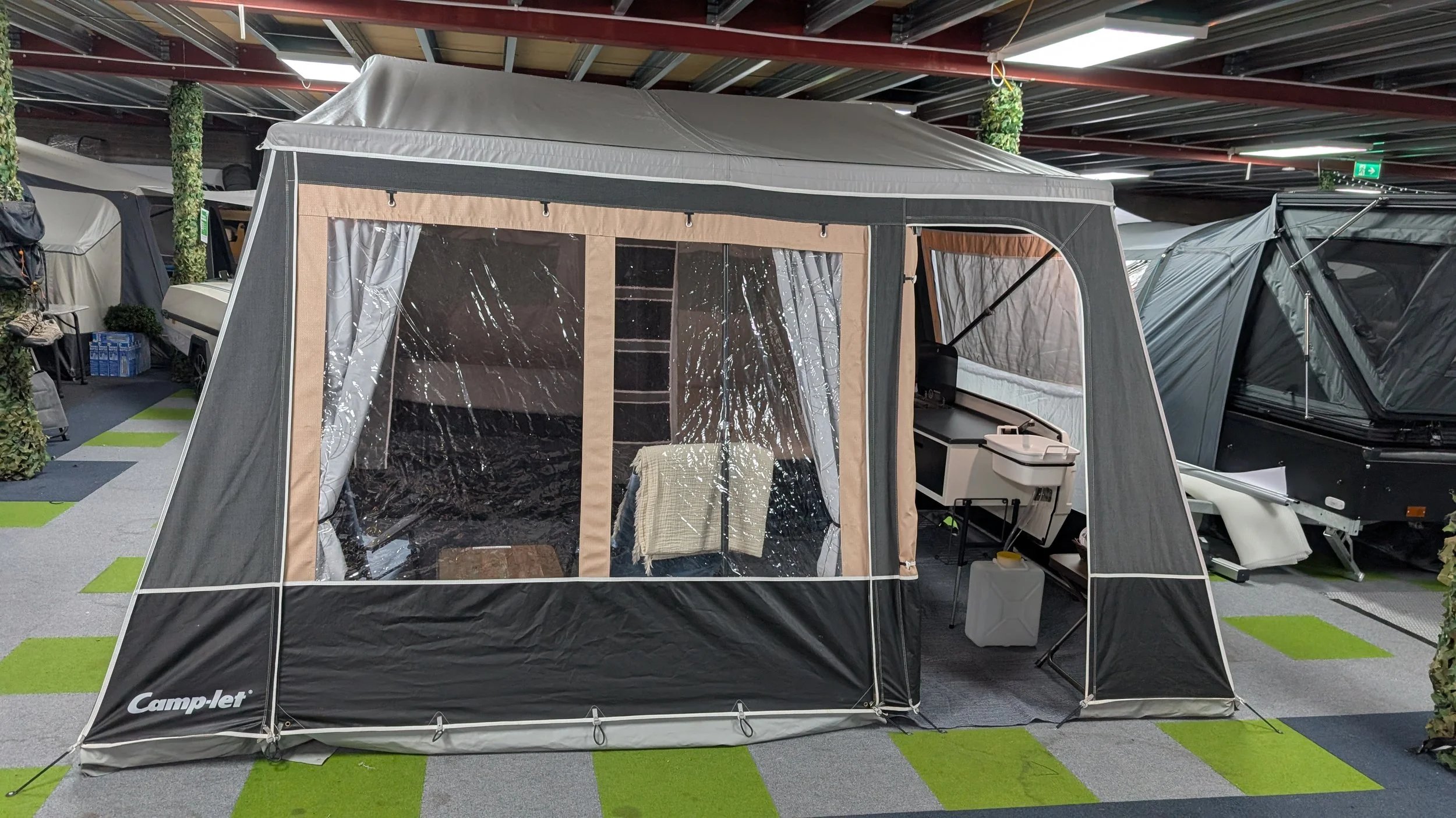 A display tent in an indoor store with transparent plastic windows and beige accents, set up on a black and green checkered floor, showing interior furniture.