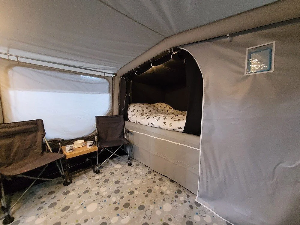 Inside a Campamster Air with a bed, two chairs, and a small table with cups. The tent has patterned flooring and a window.
