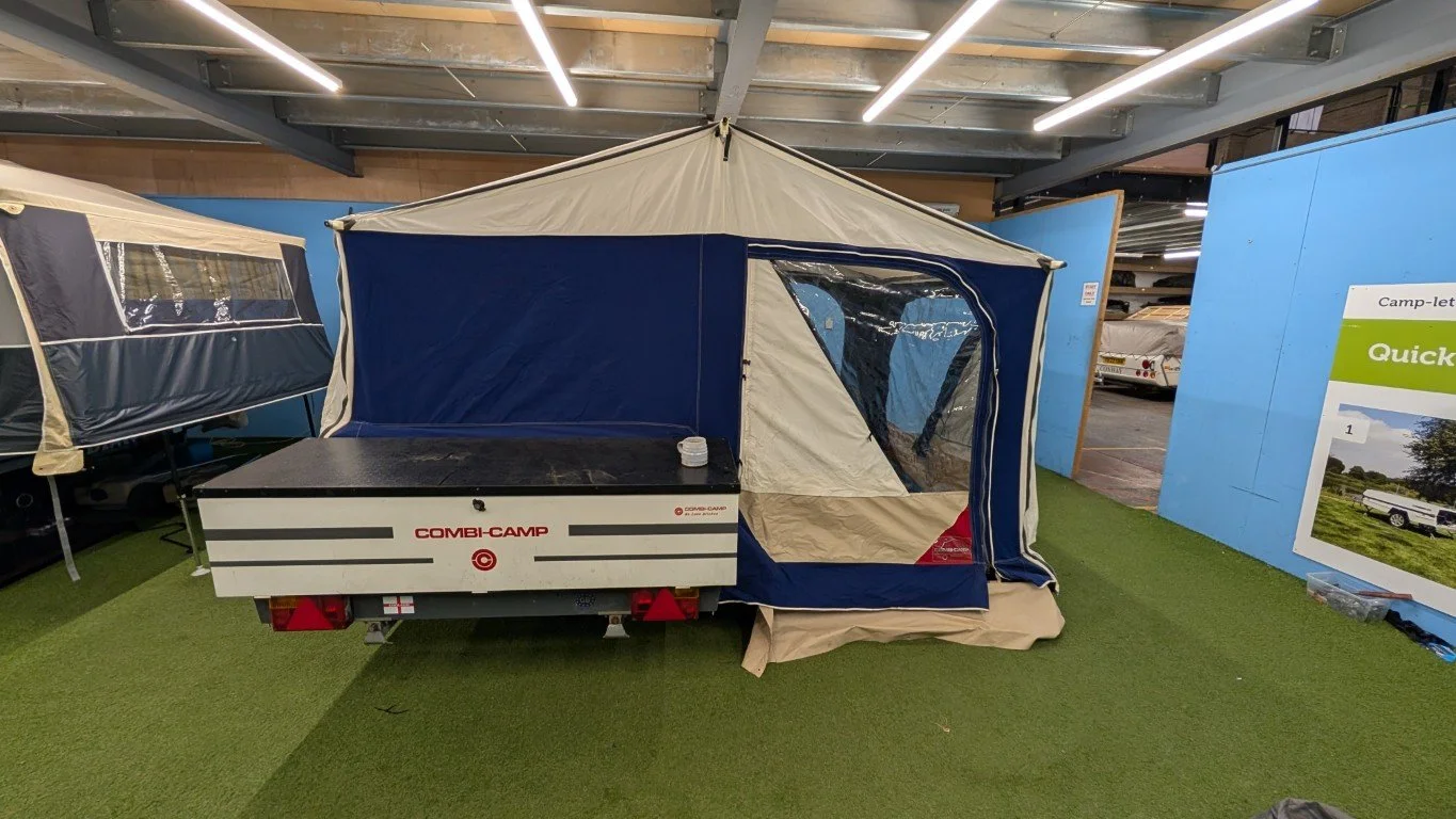 A camping tent and trailer setup display inside a building with artificial grass flooring and blue walls, featuring a sign and a few other camping accessories.