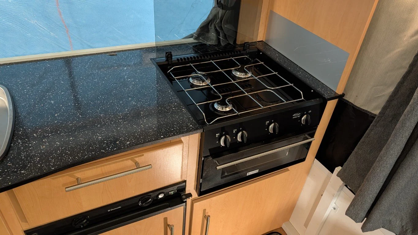 Top view of a black gas stove with four burners and an oven, set into a black speckled countertop in a kitchen. To the right of the stove is a wooden cabinet with two silver handles.