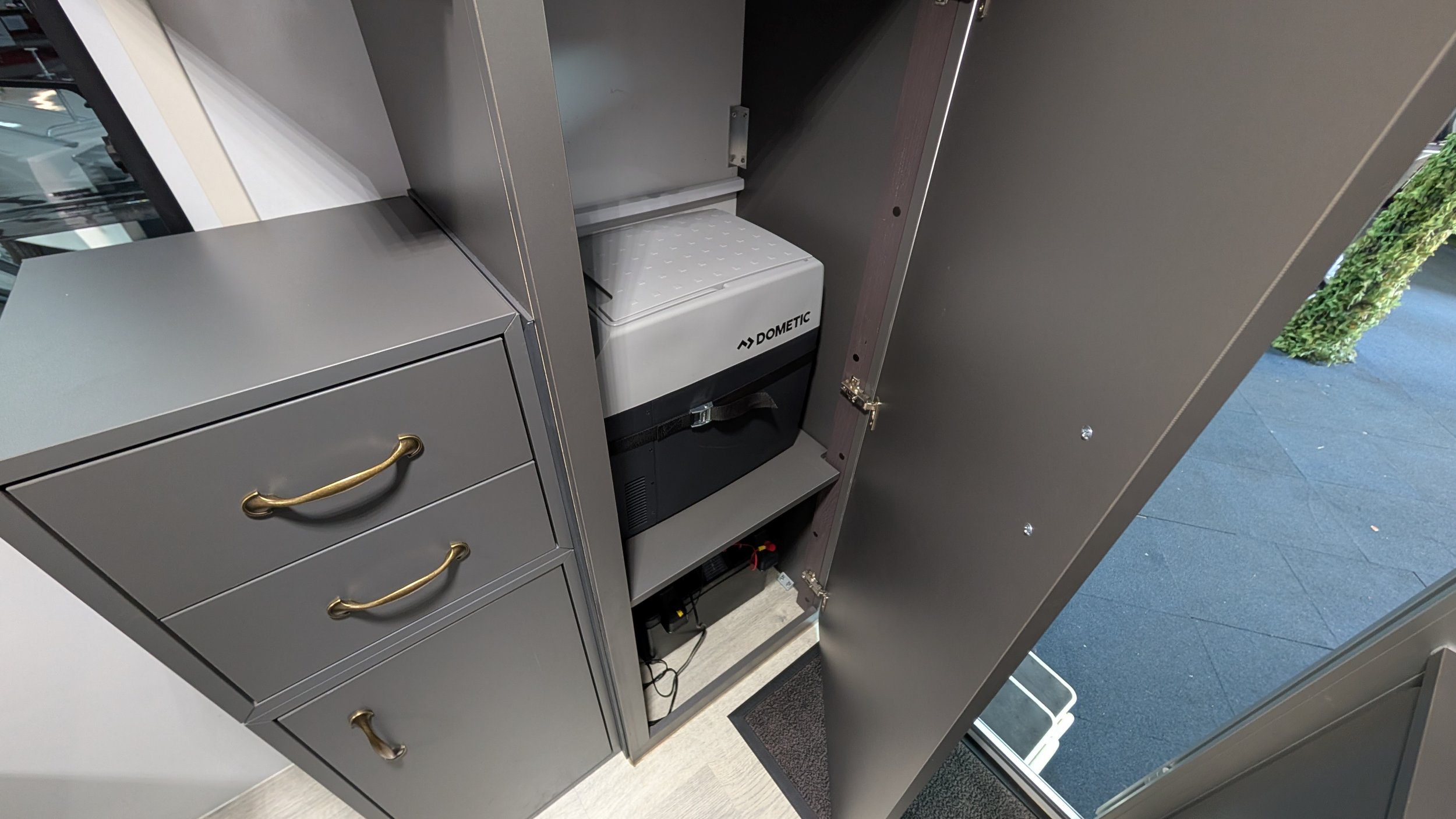 Open cabinet with Dometic refrigerator inside, located in an office or retail setting, with a gray drawer unit to the left that has gold handles. Cabinets are gray, and the refrigerator has a black base and a white door. Part of the store or office e