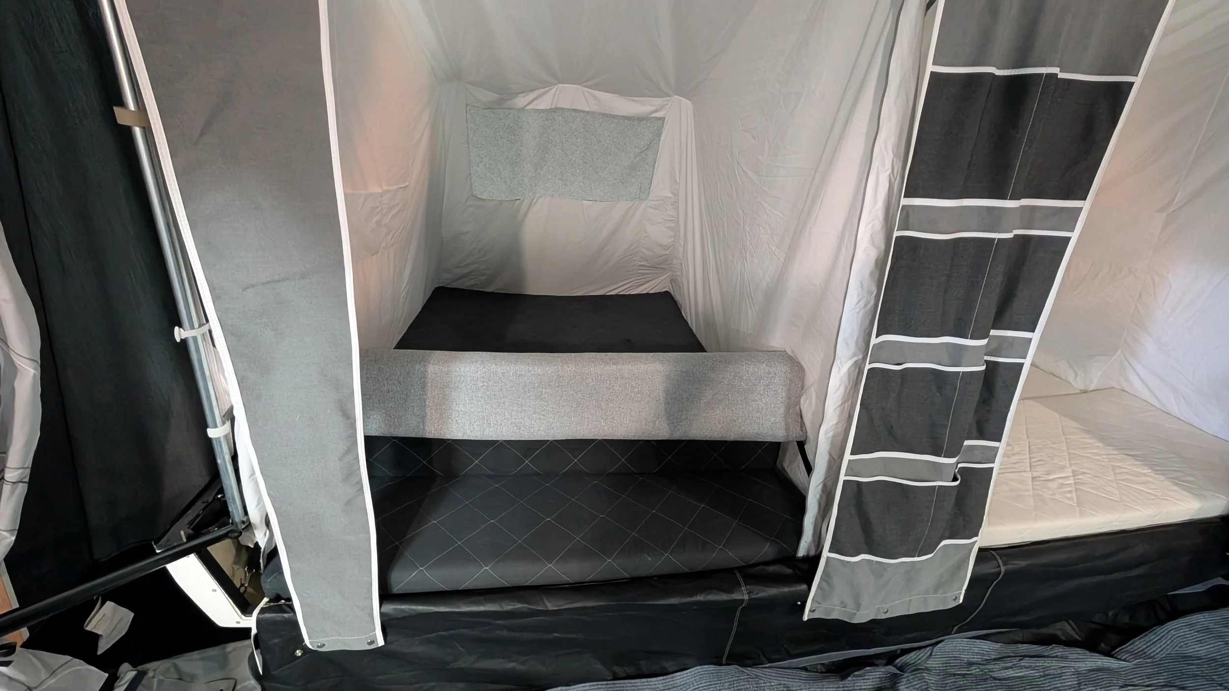 A portable camping or outdoor privacy tent with a fabric interior, folding walls, and a small interior space with a gray cushioned seat and a blanket on the wall. The tent has a black and gray window curtain on the right side.