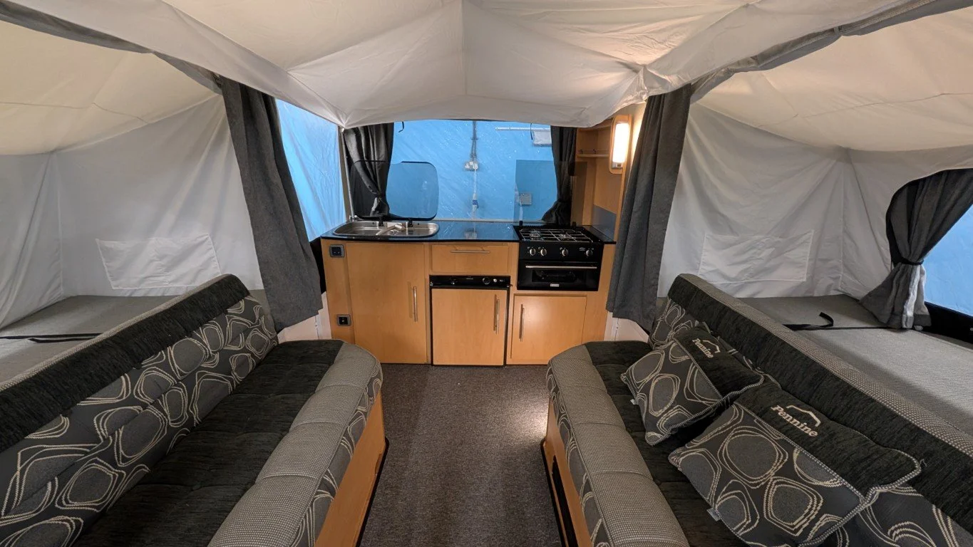 Interior of a camping tent with two black and gray sofas, a small kitchen area with a sink, stove, and wooden cabinets, and windows with black curtains.