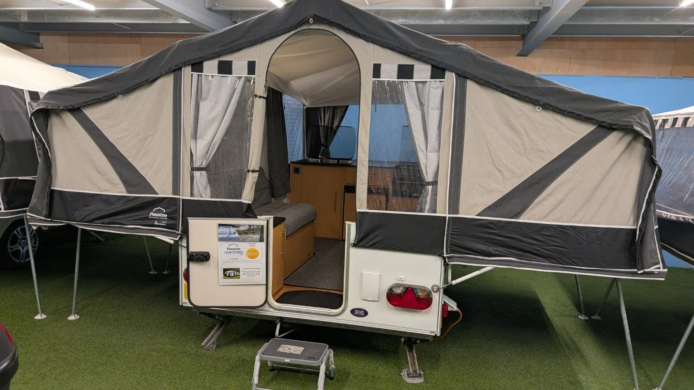 A pop-up camping trailer with its canvas cover extended, showing an interior with a bench seat, drawers, and a small TV, set up indoors on green artificial turf.