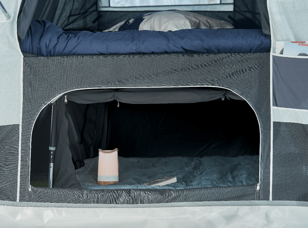 Interior of a Jamet camping tent with a lofted bed, a sleeping bag, a pillow, and a storage pocket on the side. Inside the tent's lower area, there is a portable speaker and a book on the floor.