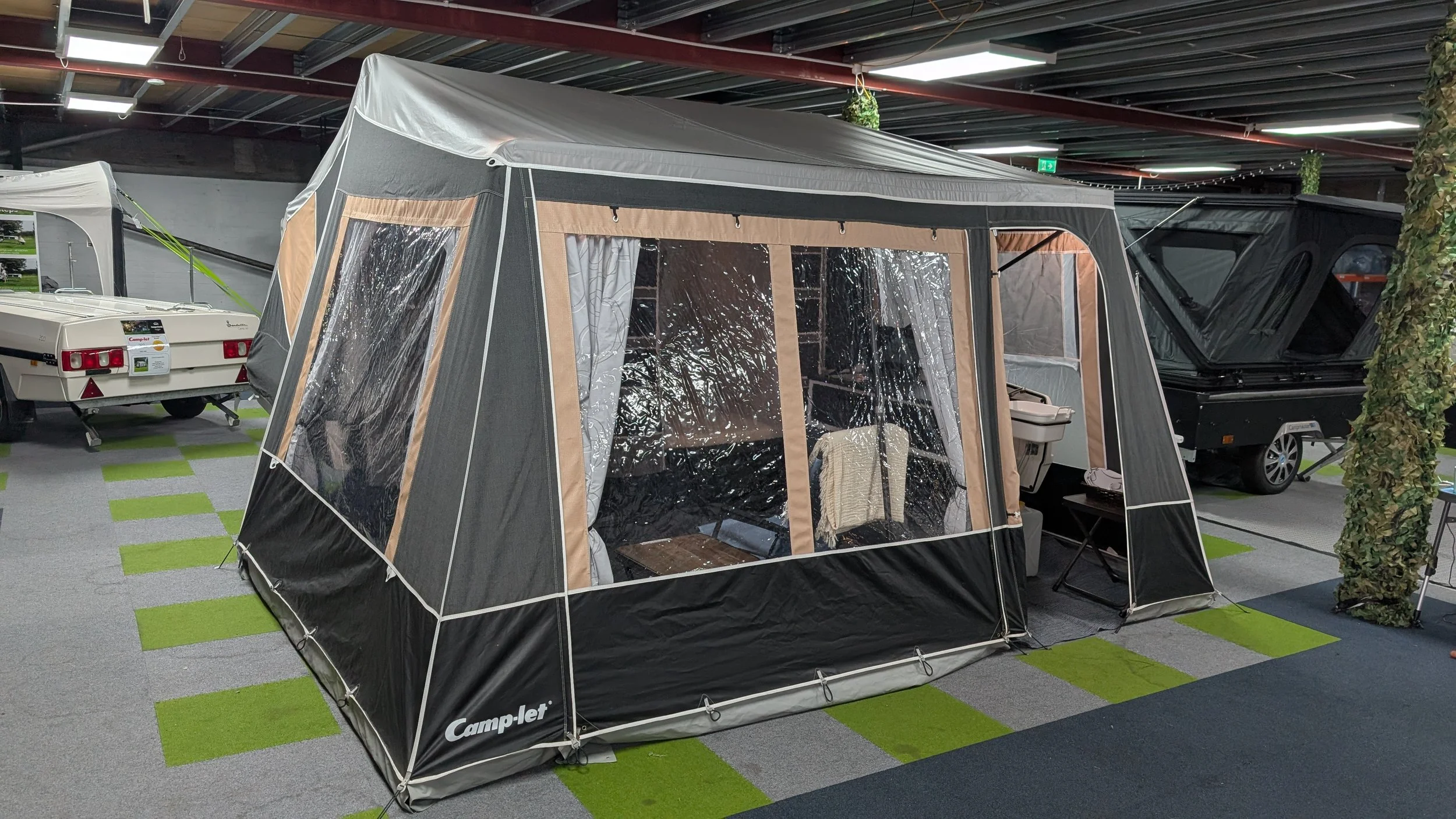 Indoor display of camping and travel trailers, with a large black and beige tent-style awning set up in the center, accompanied by a small white camper trailer and another black trailer, with green and gray checkered flooring and ceiling lights.