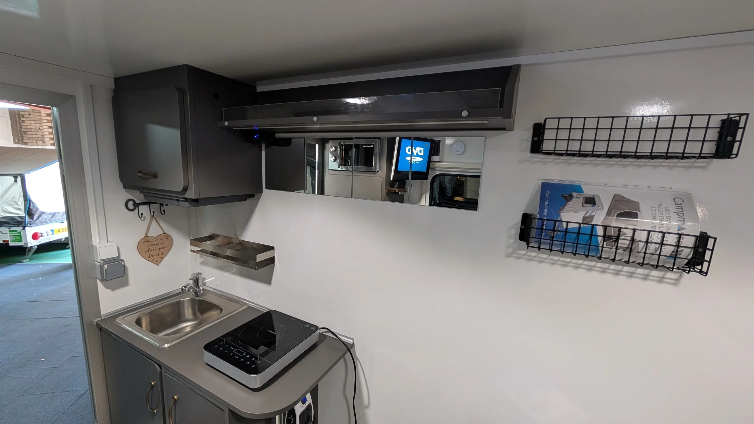 Small kitchen area with a stainless steel sink, a black single-burner stove, gray cabinets, and a mirror on the wall. There is a black wire rack holding a box and some sheets of paper. A small heart-shaped sign hangs on the wall.