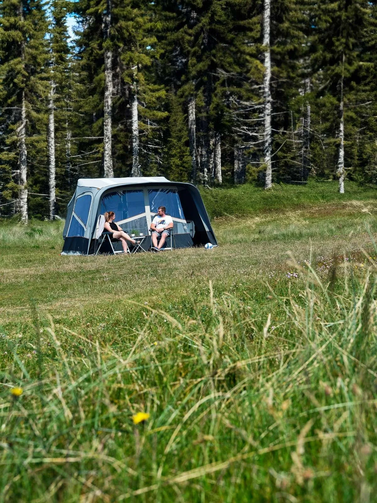 Two people sitting outside their tent in a grassy field near tall trees, having a conversation on chairs.