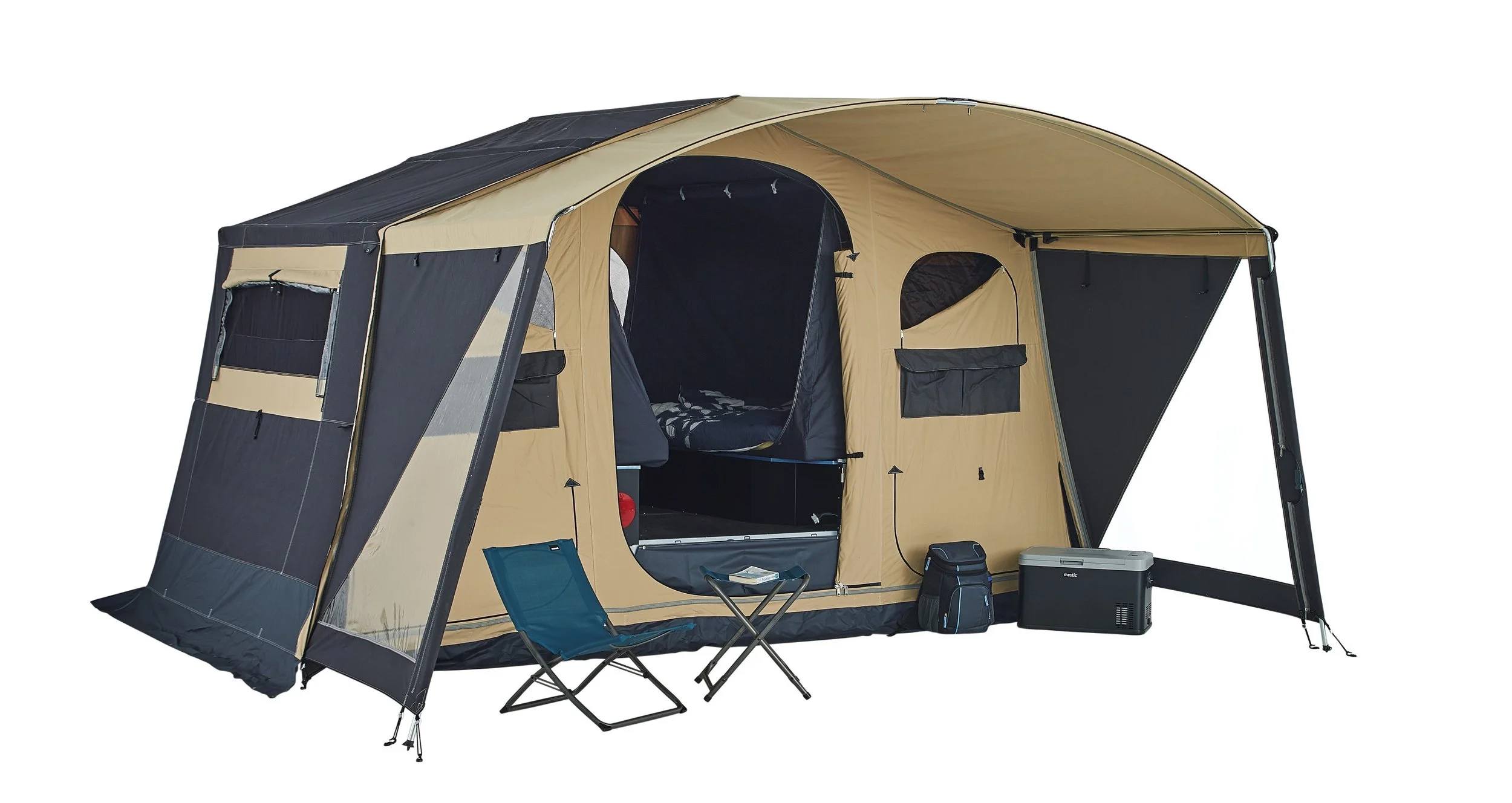 A beige and black Trigano Galleon camping tent with two windows and a large main entrance, set up with a blue portable chair, a small foldable table, a backpack, and a portable cooler, isolated on a white background.