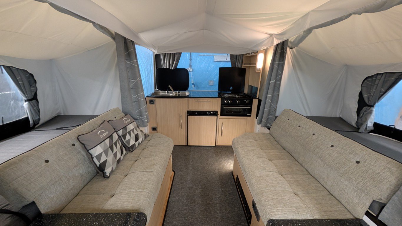 Interior of a camper tent with two beige sofas, small kitchen area with a sink, stove, and mini fridge, and windows with gray curtains.