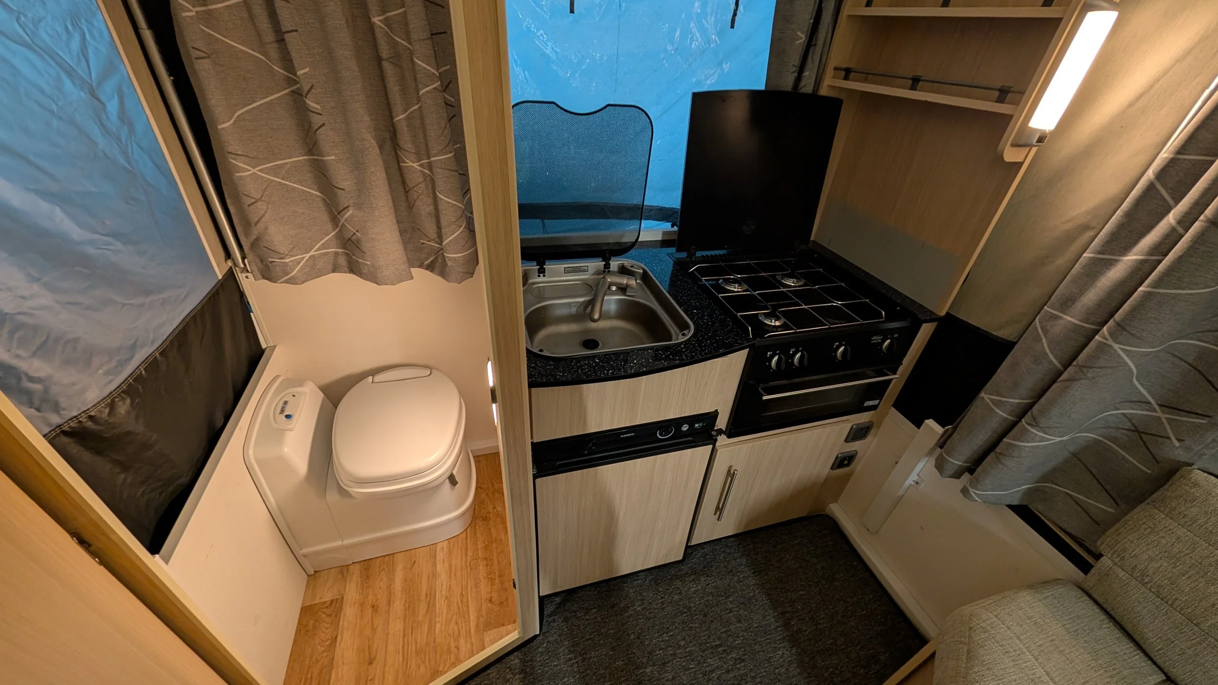 Interior view of a compact RV kitchen and bathroom area, featuring a small sink, stovetop, and toilet, with curtains and wood cabinetry.