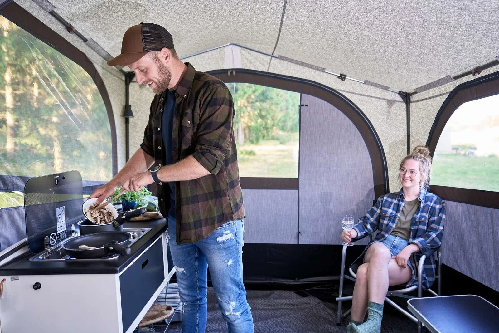 A man smiling and cooking at a portable outdoor kitchen inside a screened camping tent, while a woman relaxed in a chair holding a glass of wine and smiling.