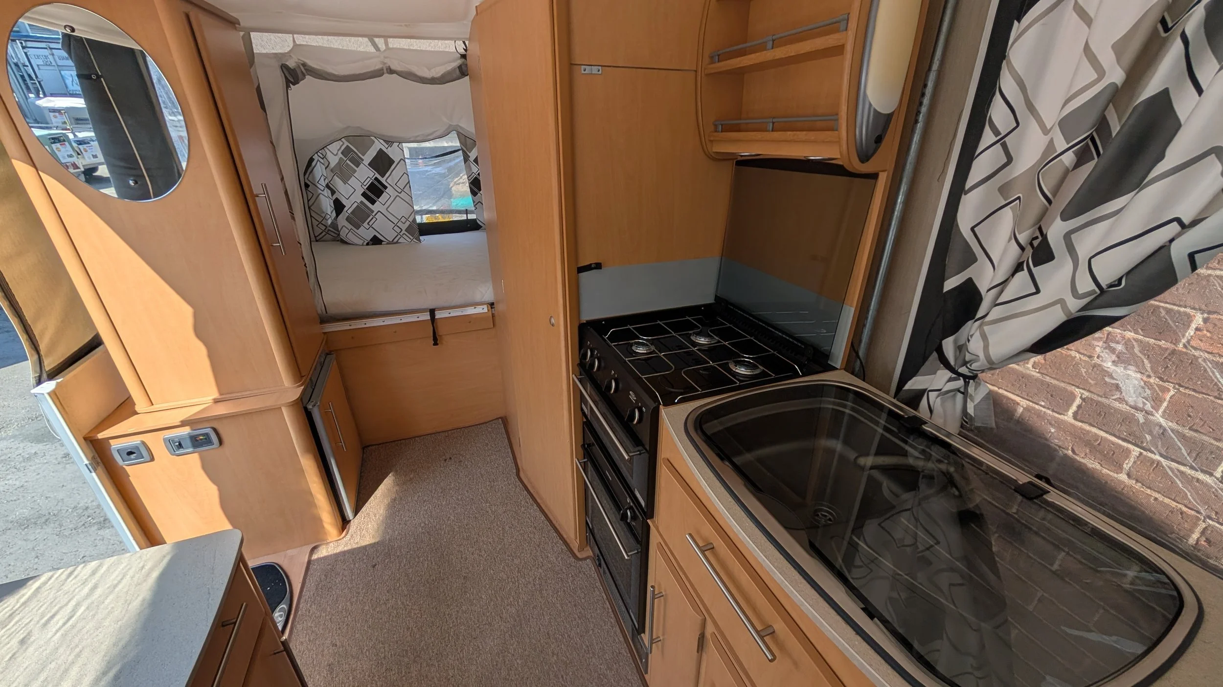 Interior of a camper van showing a stove, sink, wooden cabinets, a small bed with black and white patterned curtains, and a window with patterned curtains.