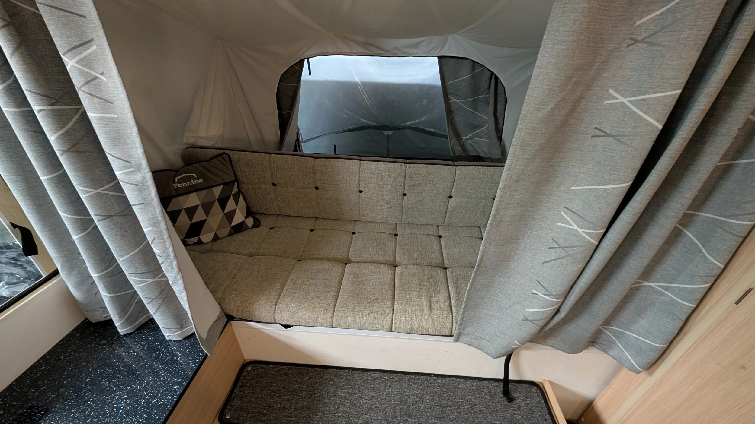 A beige cushioned booth with a window behind it, partly covered by gray curtains with white lines. There is a decorative pillow with a geometric pattern and the word 'Pennine' on it, and part of a wooden table in front.