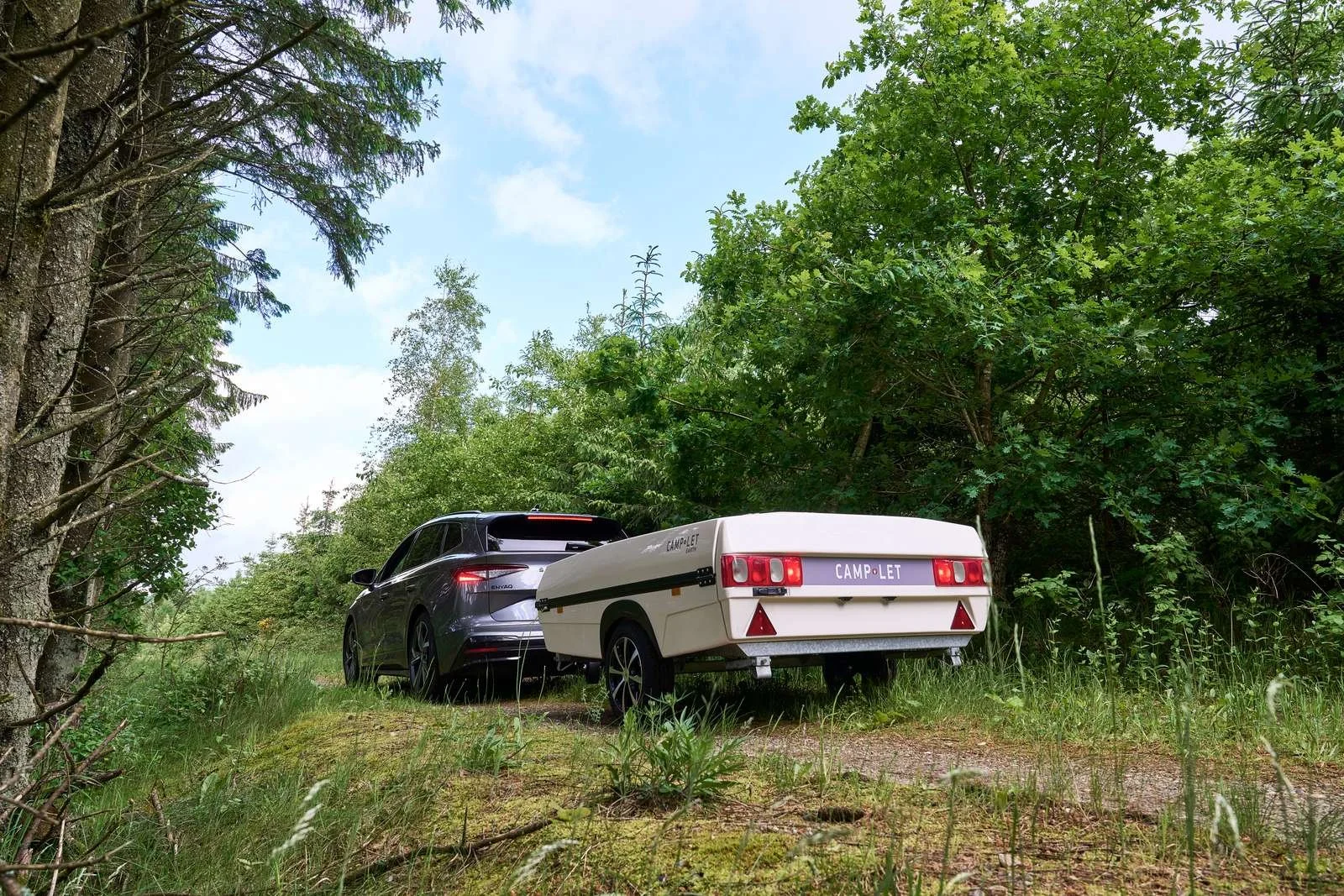 A black SUV towing a white camper trailer with the word 'CAMP•LET' on the back, parked on a grassy trail surrounded by green trees under a partly cloudy sky.