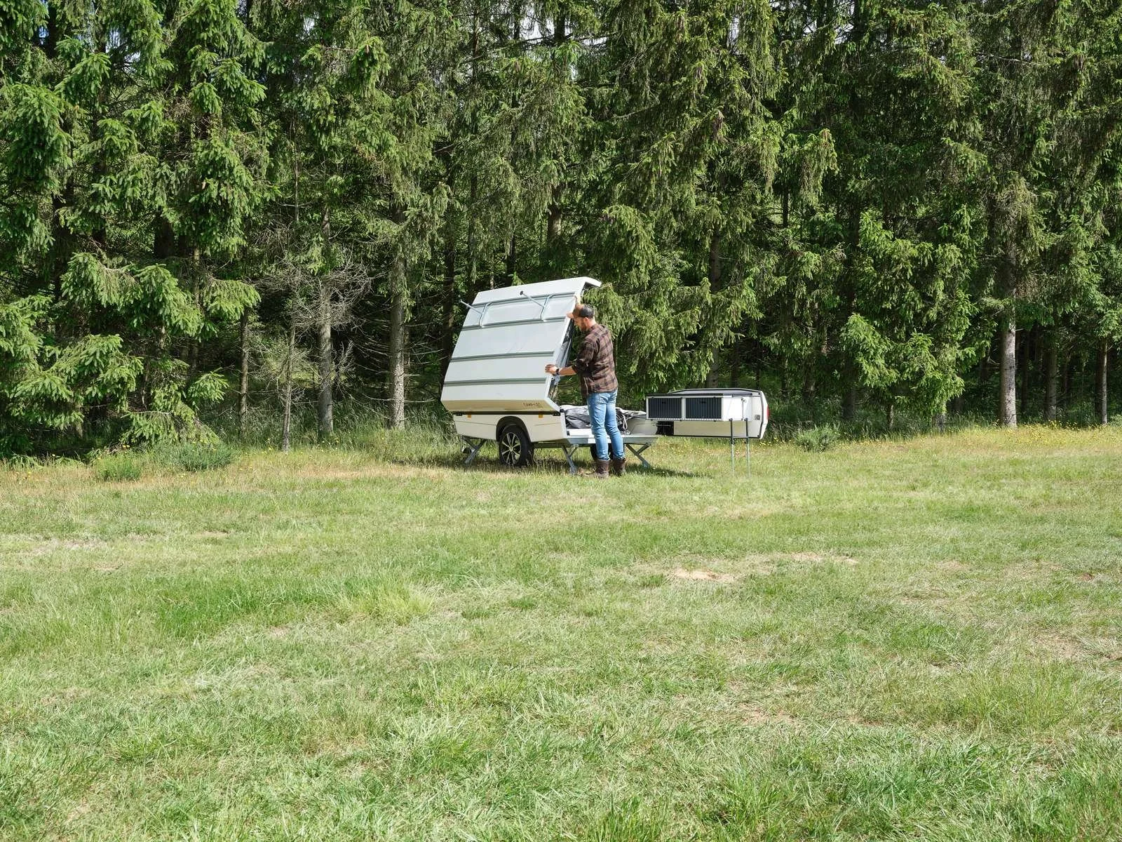 A person standing outside next to a Camp-let  with an open hatch, in a grassy field bordered by trees.