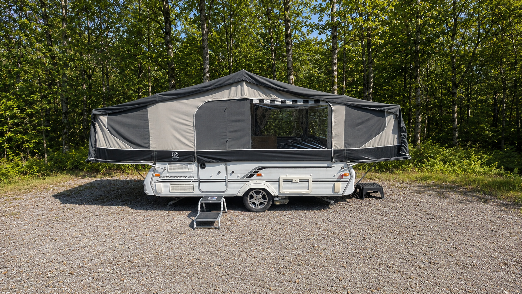 Travel trailer with pop-up tent extension parked on a gravel surface, surrounded by green trees.