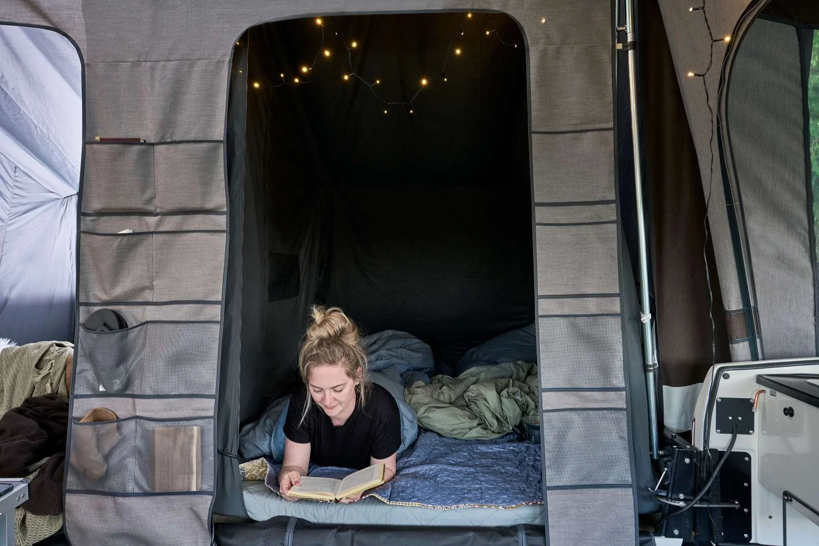 Woman lying on her stomach inside a camping tent, reading a book, with bedding and pillows around her, and string lights hanging from the ceiling.