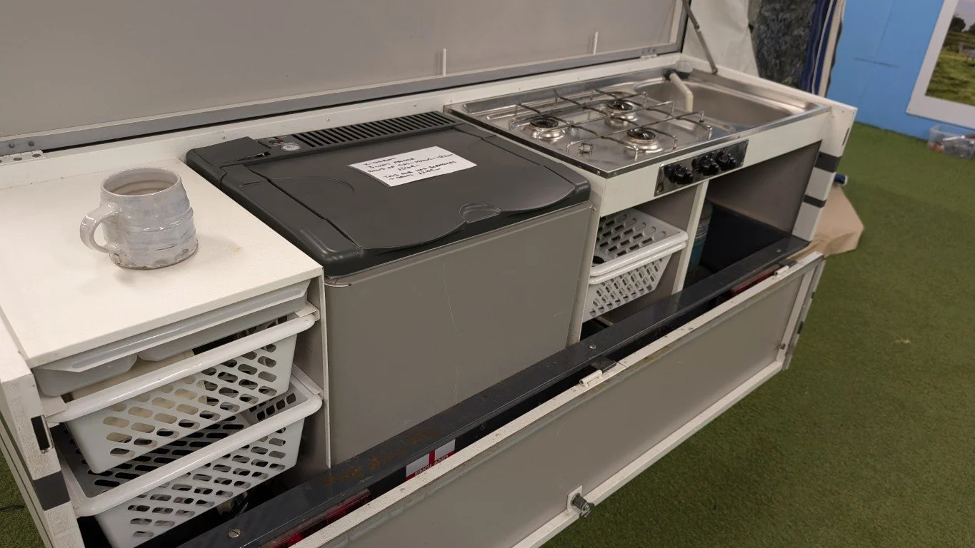 Portable outdoor kitchenette with a small stove, countertop, and storage baskets, set on artificial grass.