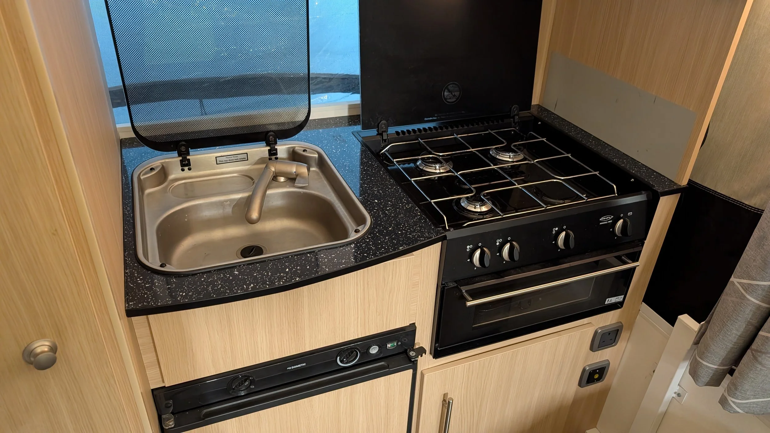 A compact kitchen area with a metallic sink with a leaf-shaped faucet, a black speckled countertop, a four-burner stove with control knobs, and a wooden cabinet surrounding the appliances.