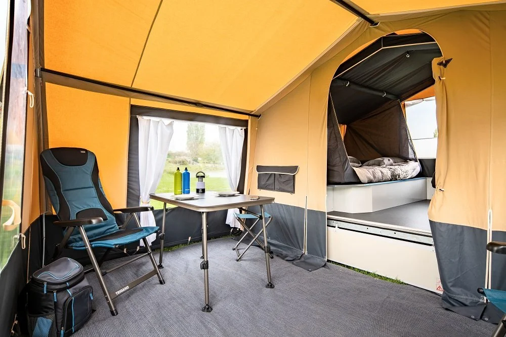 Inside a Camplair tent with a camping chair, a table with water bottles, and a view of an outdoor landscape through the window, with a sleeping area separate from the main tent space.