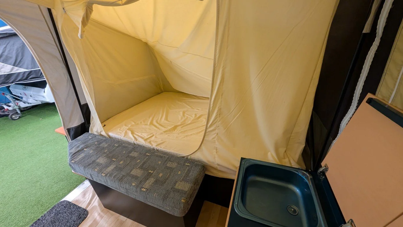 Inside a camping camper, there is a small bed with yellow fabric curtains and a gray upholstered cushion at the foot of it. To the right, there's a small black sink built into a wooden cabinet.