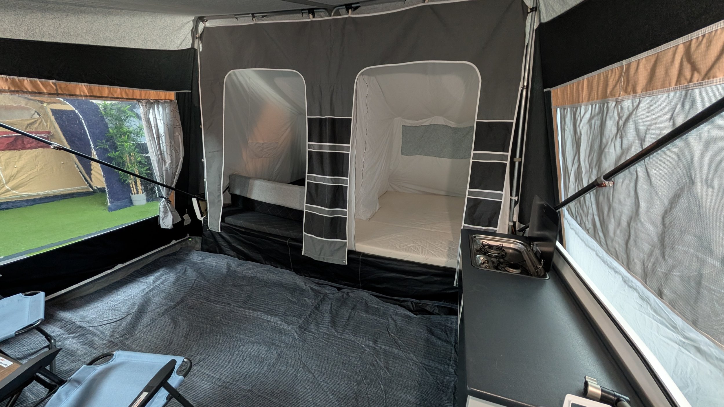 Interior of a pop-up camper with a small kitchenette, sleeping area with a bed, and tent-like fabric walls with windows showing greenery outside.