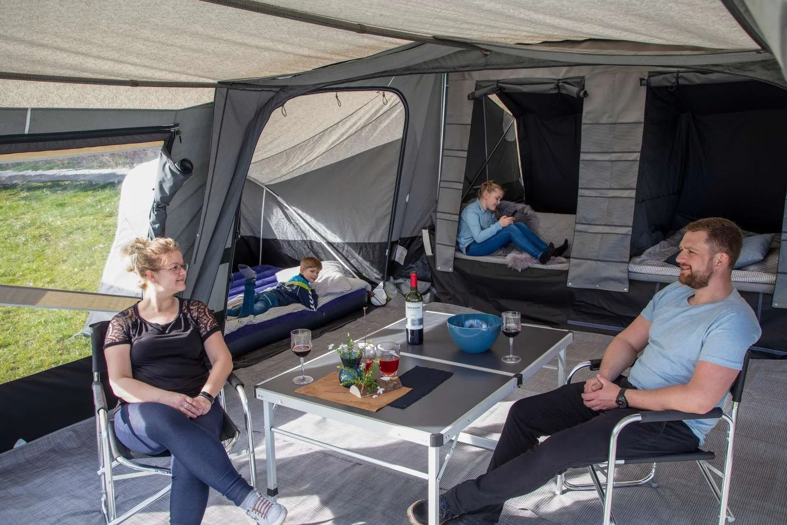 Family inside a large camping tent with two adults sitting at a table and two children in the background, one lying on a bed and the other sitting in a separate sleeping area.