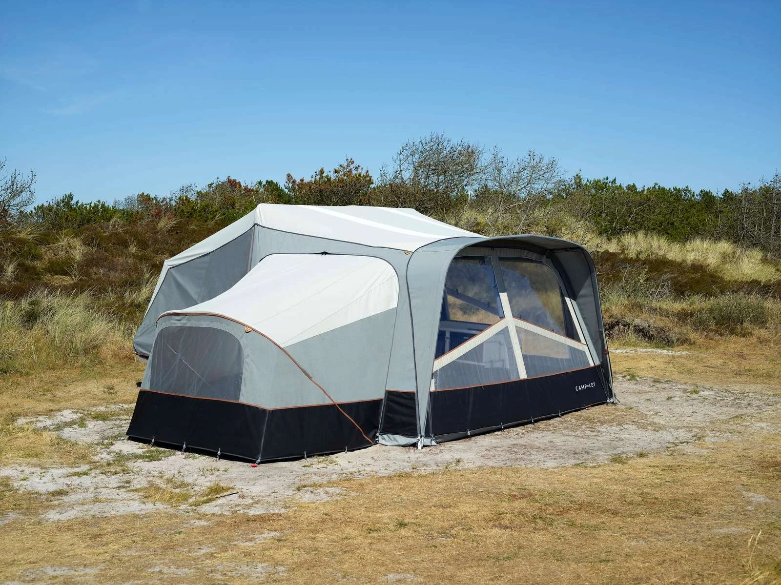 A large, modern Camp-let tent set up on a sandy and grassy area with a backdrop of bushes and small trees under a clear blue sky.
