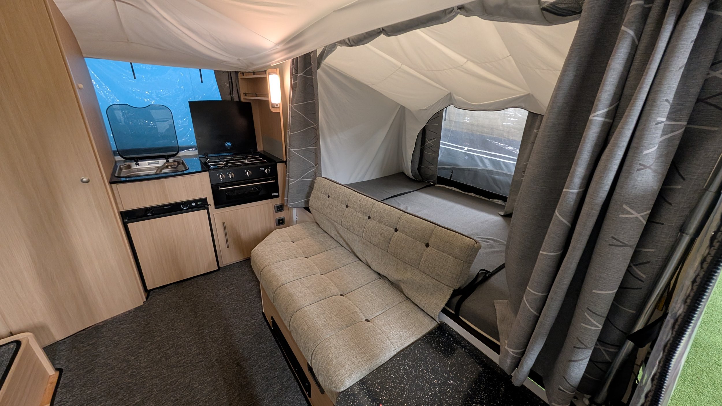 Interior of a camper with a small kitchen area, a beige couch, and sleeping area with curtains and windows.