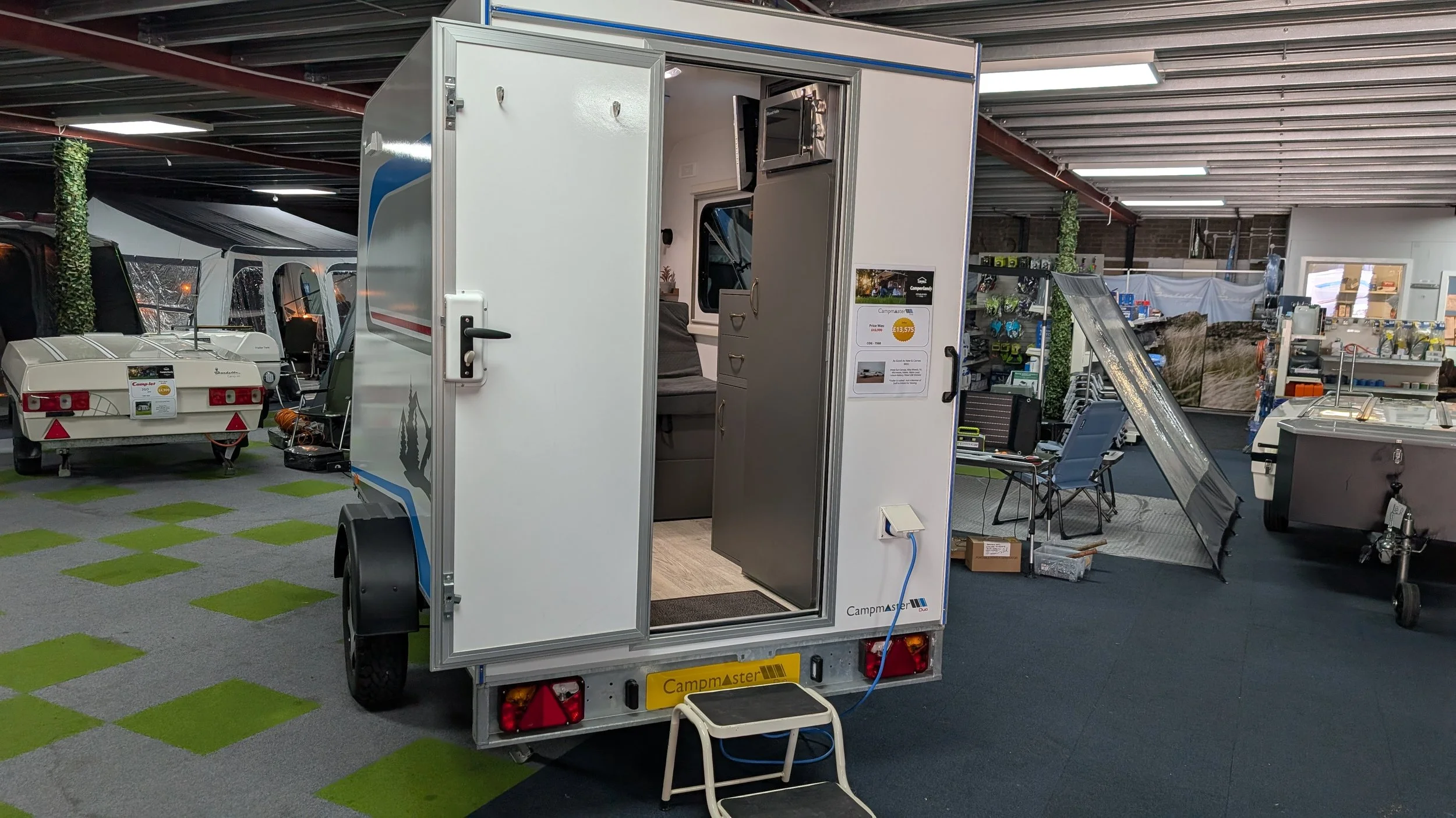 A small white camper trailer is displayed indoors, open at the door showing the interior with a couch, window, and cabinets. The surroundings include other camping equipment, trailers, and supplies in a store or showroom with ceiling lights and a par